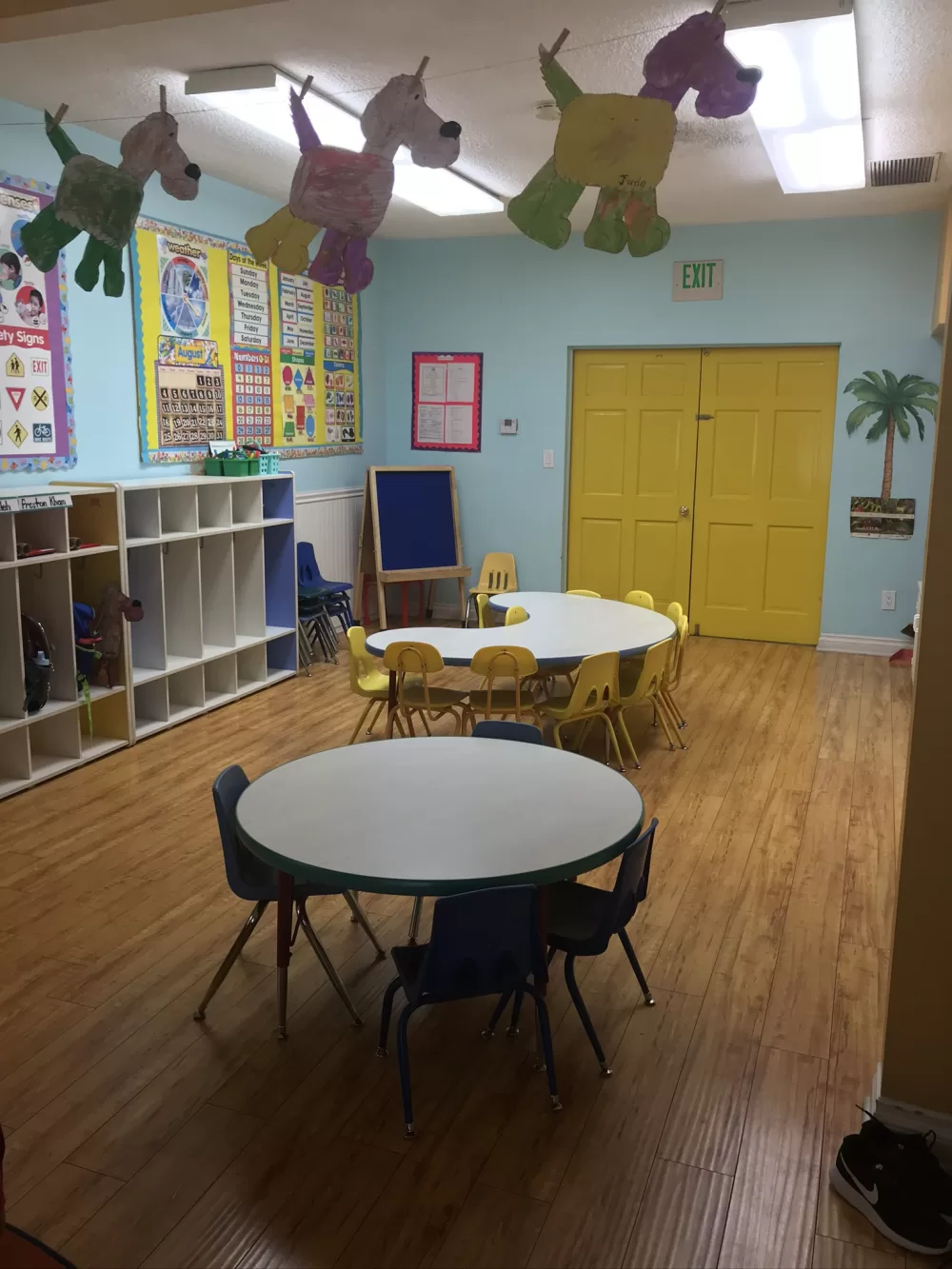 A sunlit preschool classroom with kid height tables, cubbies, and paper dogs the children made hanging overhead