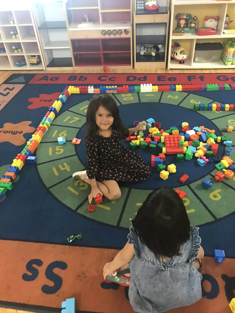 A preschooler smiles up from the counting rug while building with colorful blocks at West Hills Preschool