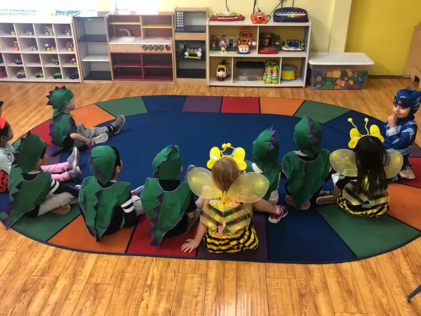 Kids in dinosaur and bumblebee costumes sit together on the rainbow reading rug