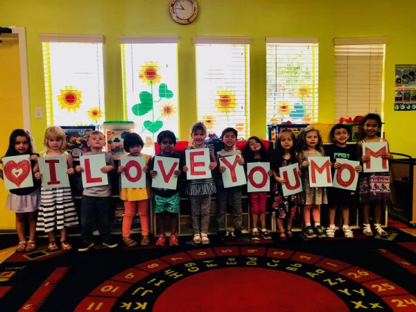 A line of preschoolers holds up painted letters spelling I love you Mom