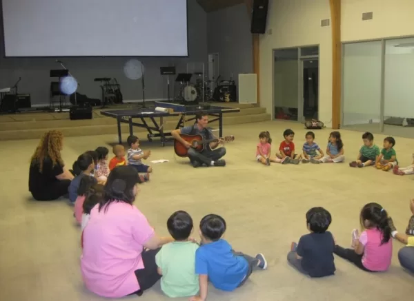 Children sit in a big circle for music time while a teacher plays live guitar